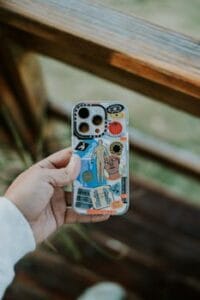 A close-up of a hand holding a creatively designed smartphone case outdoors, featuring various colorful elements. Capas para Celular.