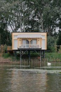 A modern wooden house on stilts by a peaceful river, surrounded by lush greenery in Hungary. Prefabricated Houses, Casas Pré-Fabricadas.