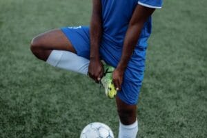 A soccer player in a blue uniform stretches on the grassy field, preparing for the game. Análise das Chuteiras.