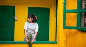 A woman stands in a bright yellow and green environment, holding a bouquet of flowers. Casa Verde e Amarela.