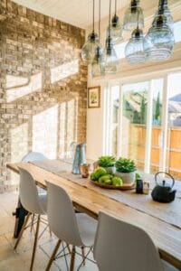 Sunlit dining room with contemporary decor and a rustic brick wall, featuring stylish lighting and a wooden table. Decoration, Decoração.