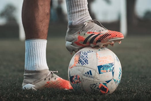 Close-up of a soccer player wearing Adidas cleats, holding a branded soccer ball on grass. Football boot, Chuteira.