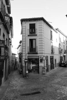 Black and white photo of a quaint street corner in Toledo, Spain, showcasing historic architecture. Casa.