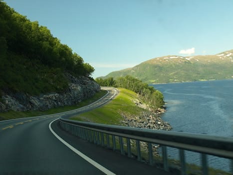 Breathtaking view of a winding road along a Norwegian fjord, surrounded by water and lush greenery. Trip, Viagem.