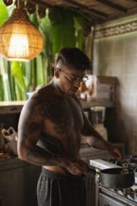 A shirtless man with tattoos cooking and stirring a pot in a rustic indoor kitchen. Fit Kitchen, Cozinha Fit.