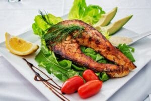 Delicious grilled salmon steak served with avocado, lettuce, cherry tomatoes, and lemon slice on a white plate. Fish food, comida, food.