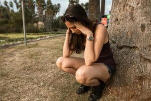 A woman kneels by a tree holding her head, expressing stress or anxiety in an outdoor park. Anxiety, Ansiedade.