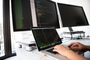 A developer writes code on a laptop in front of multiple monitors in an office setting. Technology, Tecnologia.