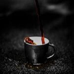 Close-up of steaming dark coffee being poured into a black cup on a dark textured surface. Rich aroma. Café, Coffee, comida, food.