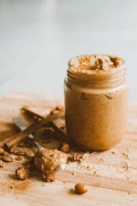 A jar of creamy peanut butter with scattered peanuts and spoon on a wooden surface. peanut food, food, comida, amendoim.